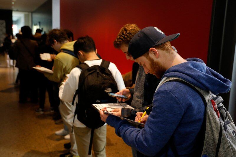 A man in a sweatshirt and backpack stands in line and write on a clipboard
