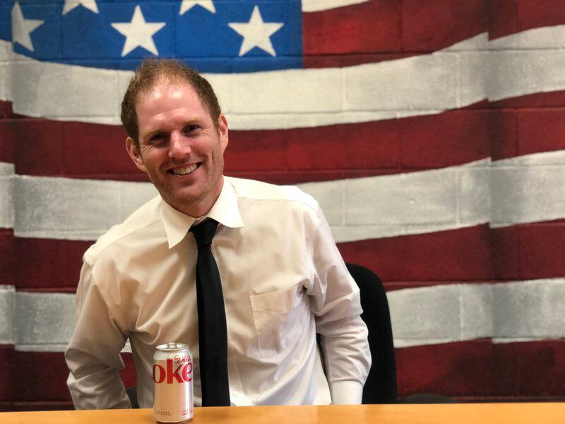 A man in a shirt and tie sits at a desk with an American flag painted on the wall behind him