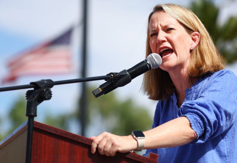 A woman speaks into a microphone outside as an American flag waves in the background