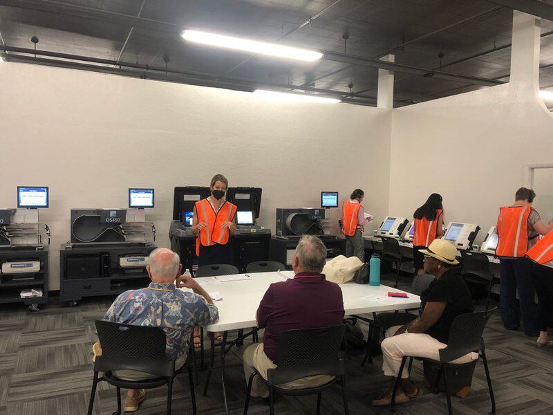 A woman in an orange vest gestures before computerized machines while three seated observers watch.