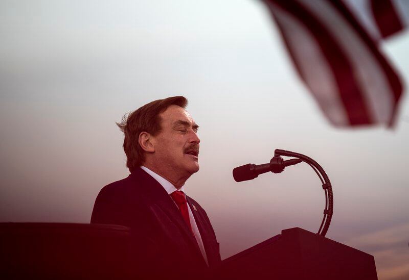 Mike Lindell’s eyes are closed while he stands outdoor at a lectern against a twilight sky