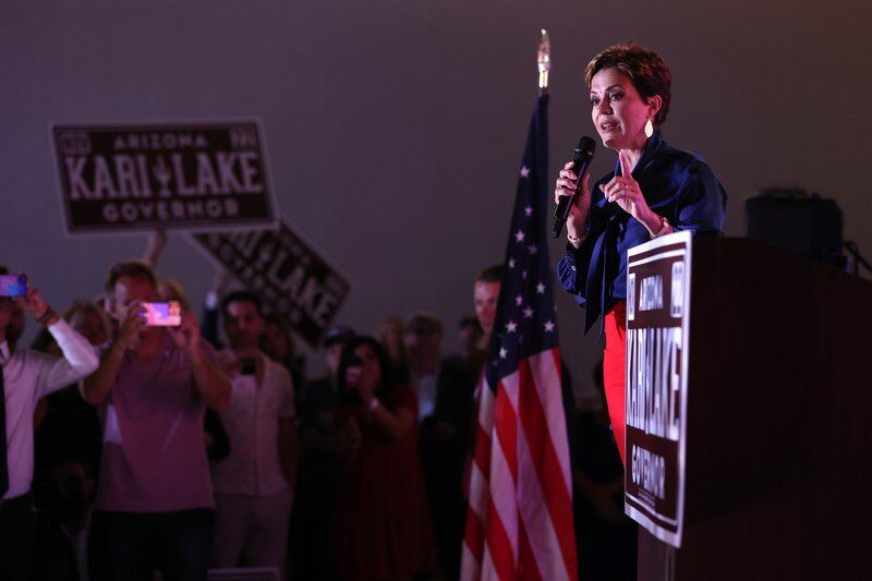 A woman speaks from a stage facing a crowd holding up signs that say “Kari Lake”