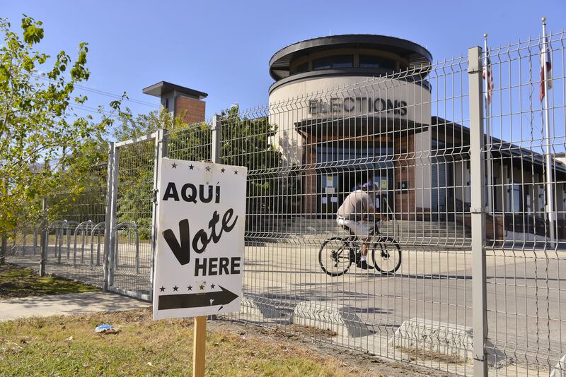A sign that reads “vote here” points to a large building in the background and a man is seen riding his bike in front of it. 