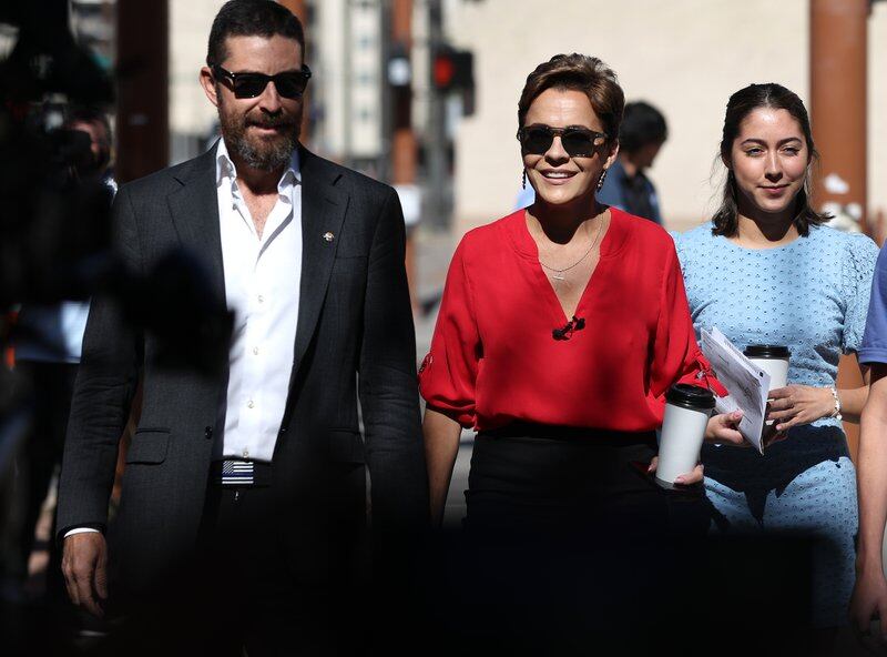 A bearded man in a dark suit and sunglasses holds the hand of a woman wearing a red blouse and sunglasses and holding a coffee cup as they walk.