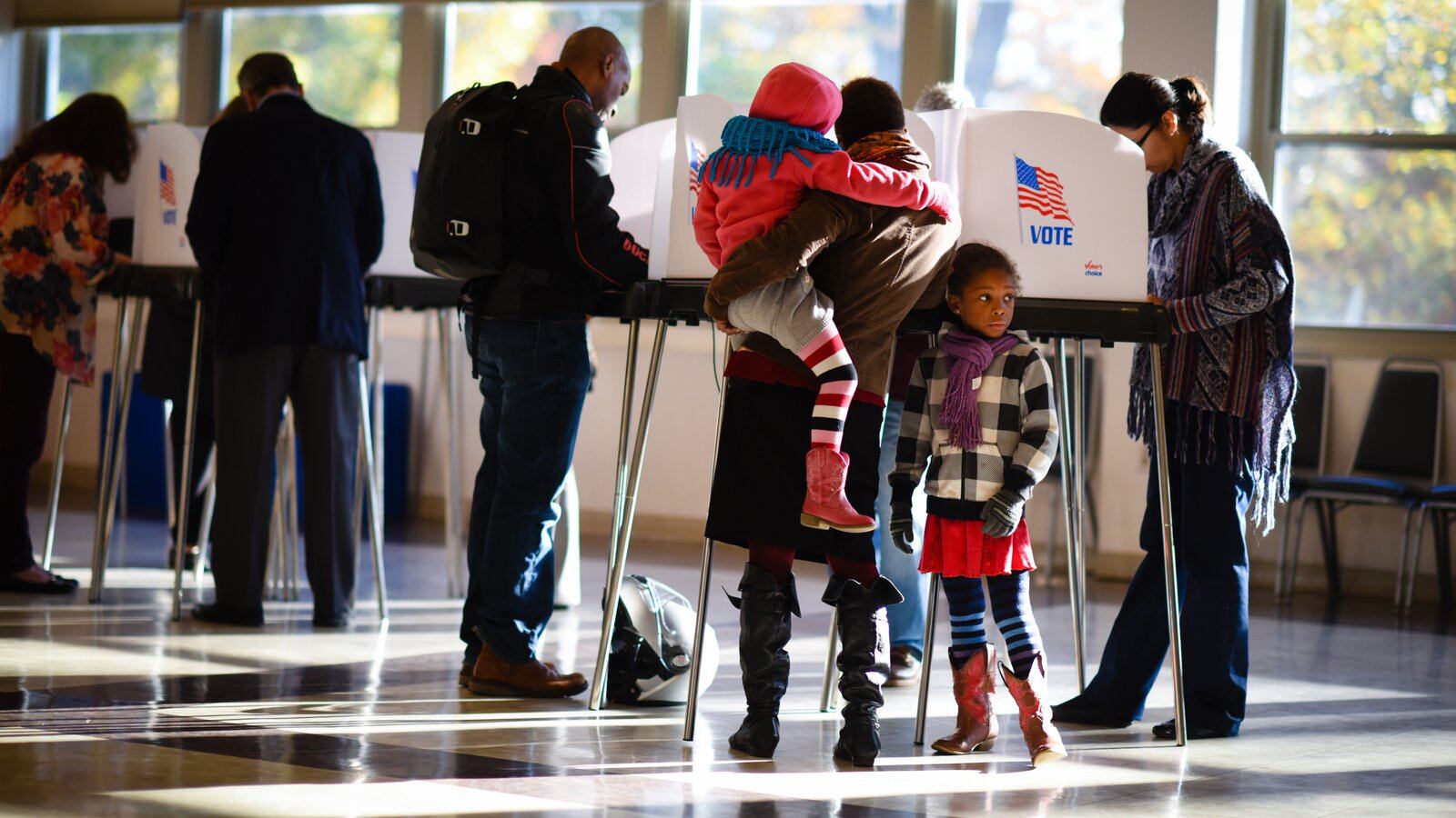 A woman holding a small child stands at a row of polling place booths, along with other voters, as a girl looks on.