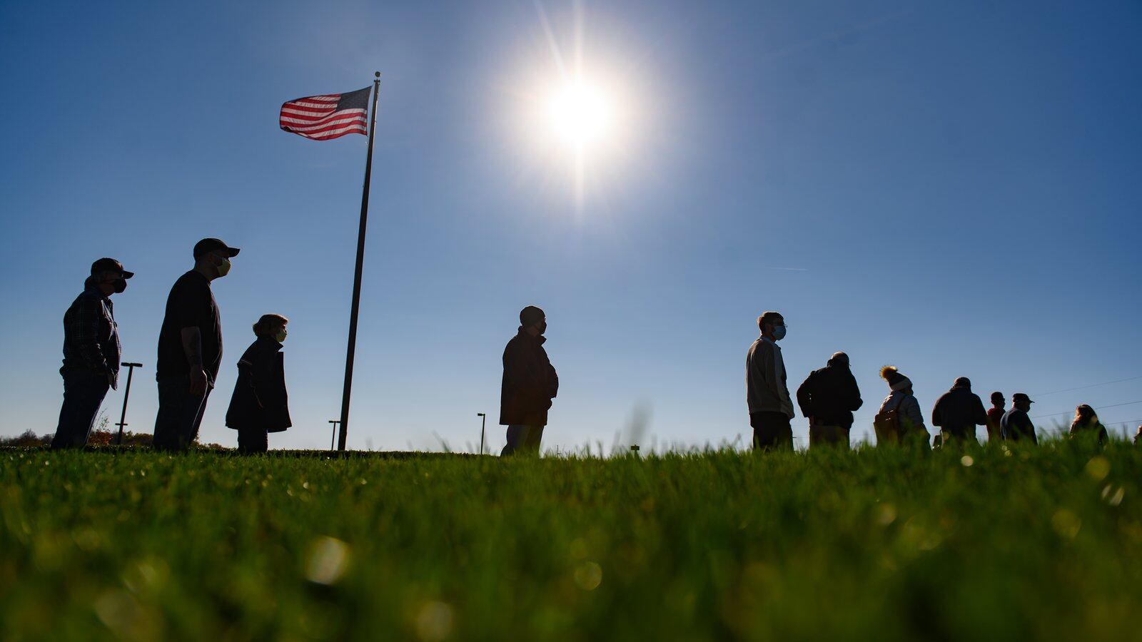 A row of people in silhouette standing on a grassy hill with a U.S. flag waving in the background