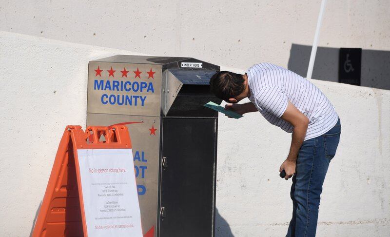 A man bends down to look into the opening of a silver drop box on a sidewalk