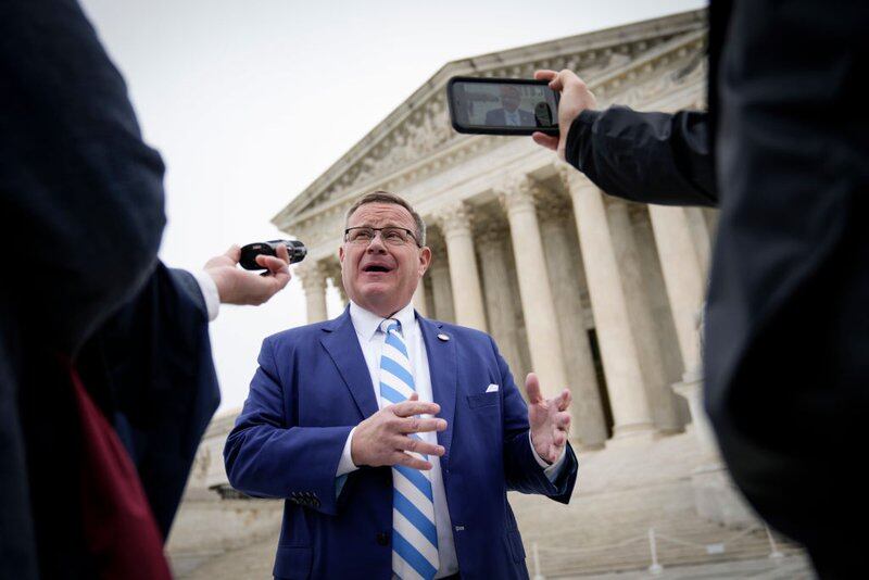A man with glasses wearing a striped blue-and-white tie and blue blazer gestures while speaking outside at the foot of a flight of steps leading up to a building with classical columns.