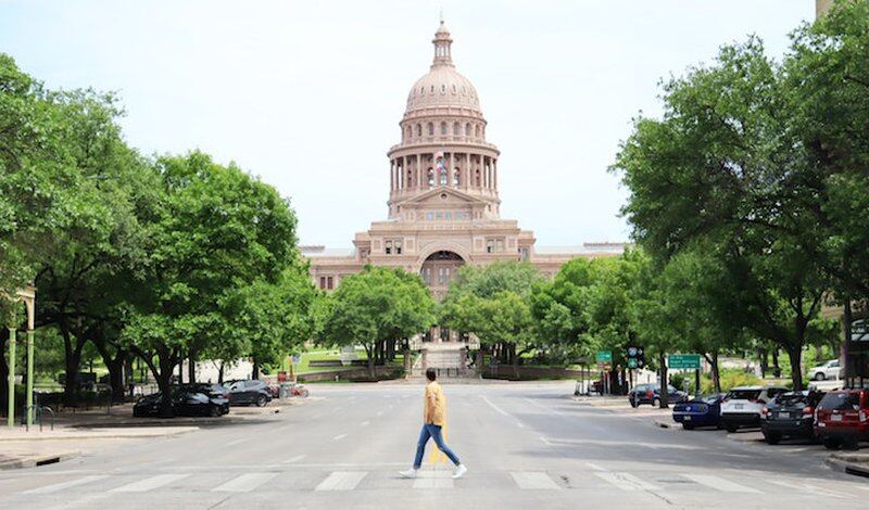 A man uses a crosswalk to cross a street and in the background is a statehouse building