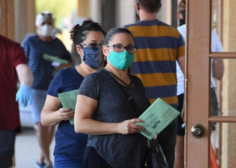 Two people with face masks wait outside door of polling place with green ballot envelopes in their hands.