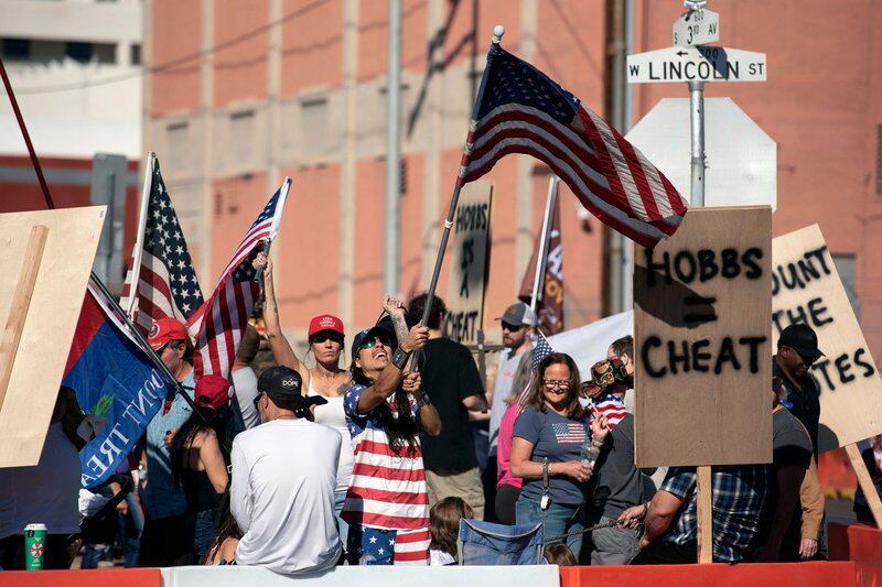 People crowd together, waving American flags and handmade signs, outside a red brick building.