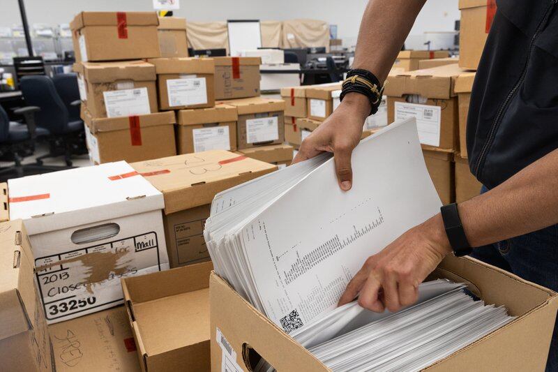 A man reaches into a cardboard file box of ballots
