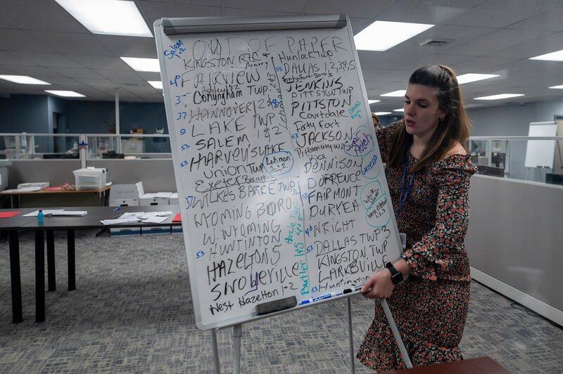 A woman lifts a large dry-erase board covered in writing, topped by the words “Out of paper”
