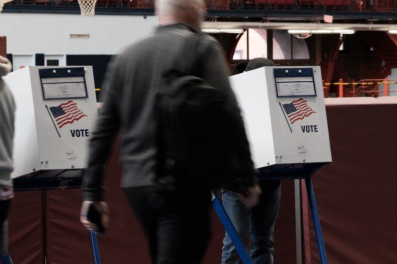 A man walks through a gymnasium past a row of voting booths, each decorated with an American flag and the word “Vote.”
