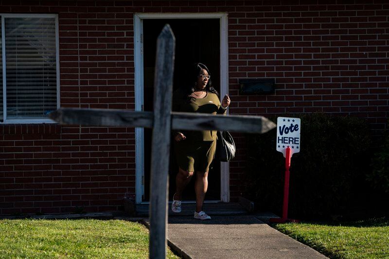 A woman walks out of a building with a “Vote Here” sign next to the door. A cross sits in the foreground of the photograph.