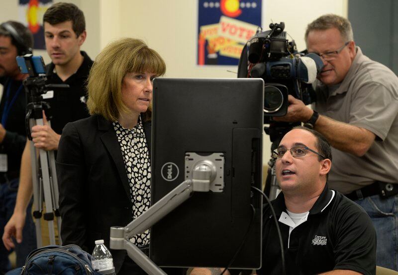 A woman stands next to a man seated before a computer screen as they both look at it, with a man holding a television camera in the back, capturing the scene.