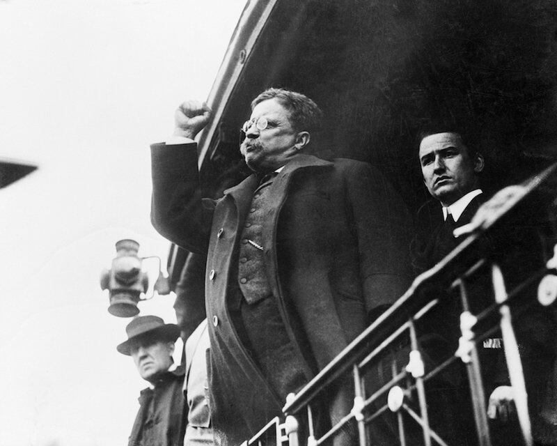 Black and white photograph of President Roosevelt standing at a railing with his fist raised aggressively.