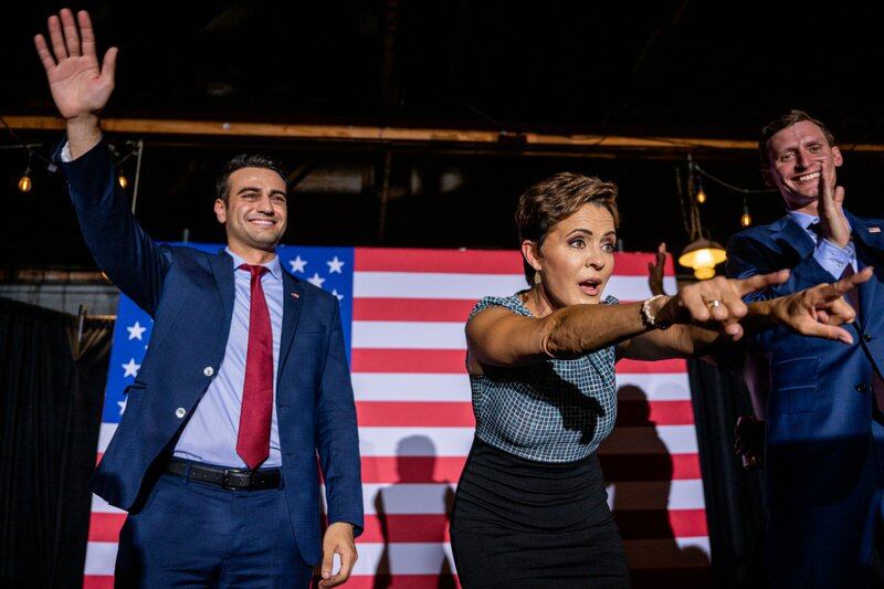 A woman and two men point and wave from a stage, an American flag hung behind them