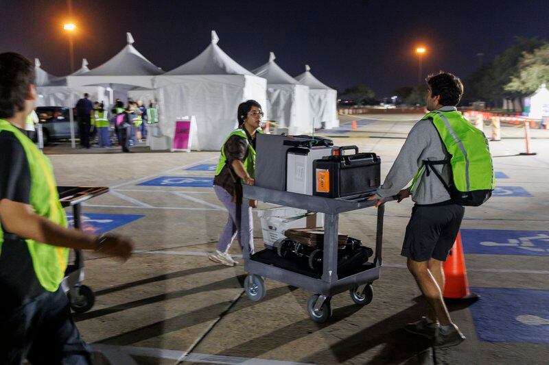 Two workers in yellow vests wheel a cart of election supplies across a parking lot at night.
