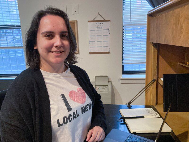 Woman sitting at a desk wearing a T-shirt that reads “I love local news”