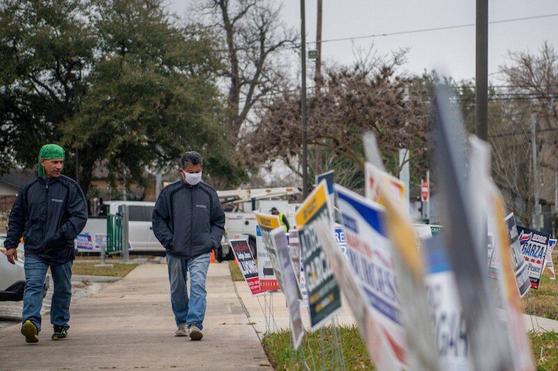 Two men walk down a sidewalk lined on one side with dozens of campaign signs, shown as a blur.