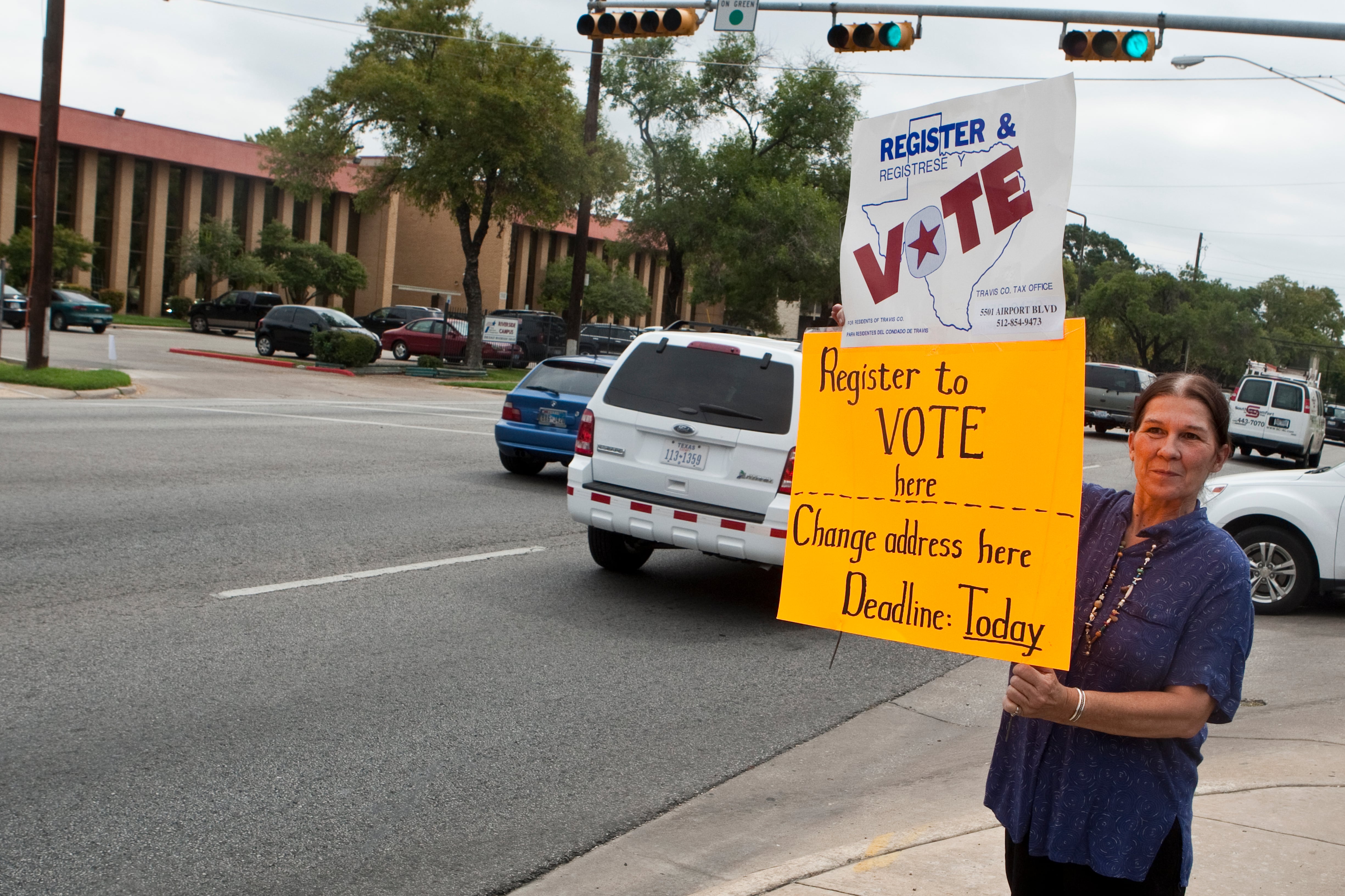 A woman stands at a street corner with signs reading, “Register to vote here,” and “Change address here. Deadline today.”