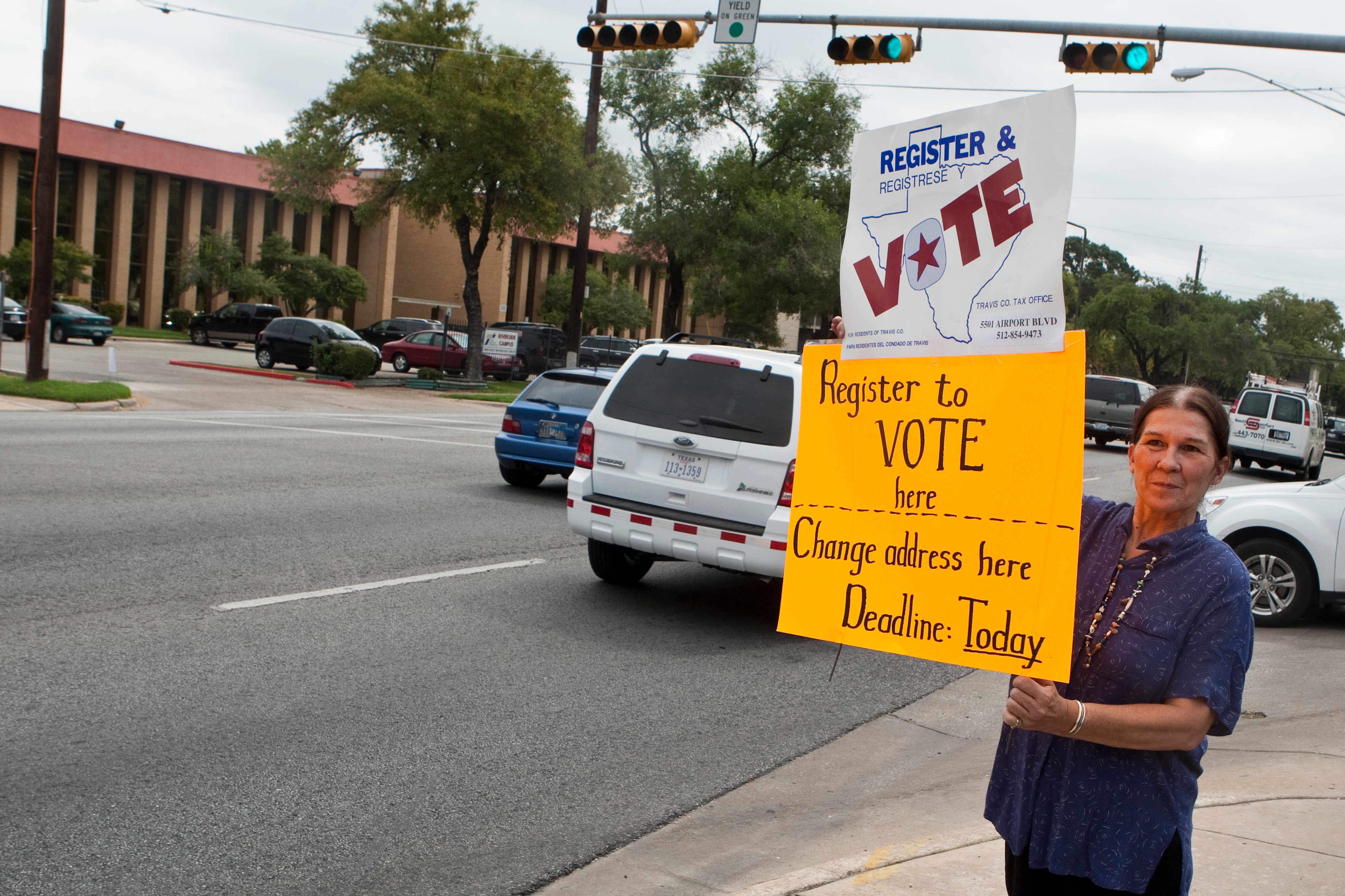 A woman stands at a street corner with signs reading, “Register to vote here,” and “Change address here. Deadline today.”