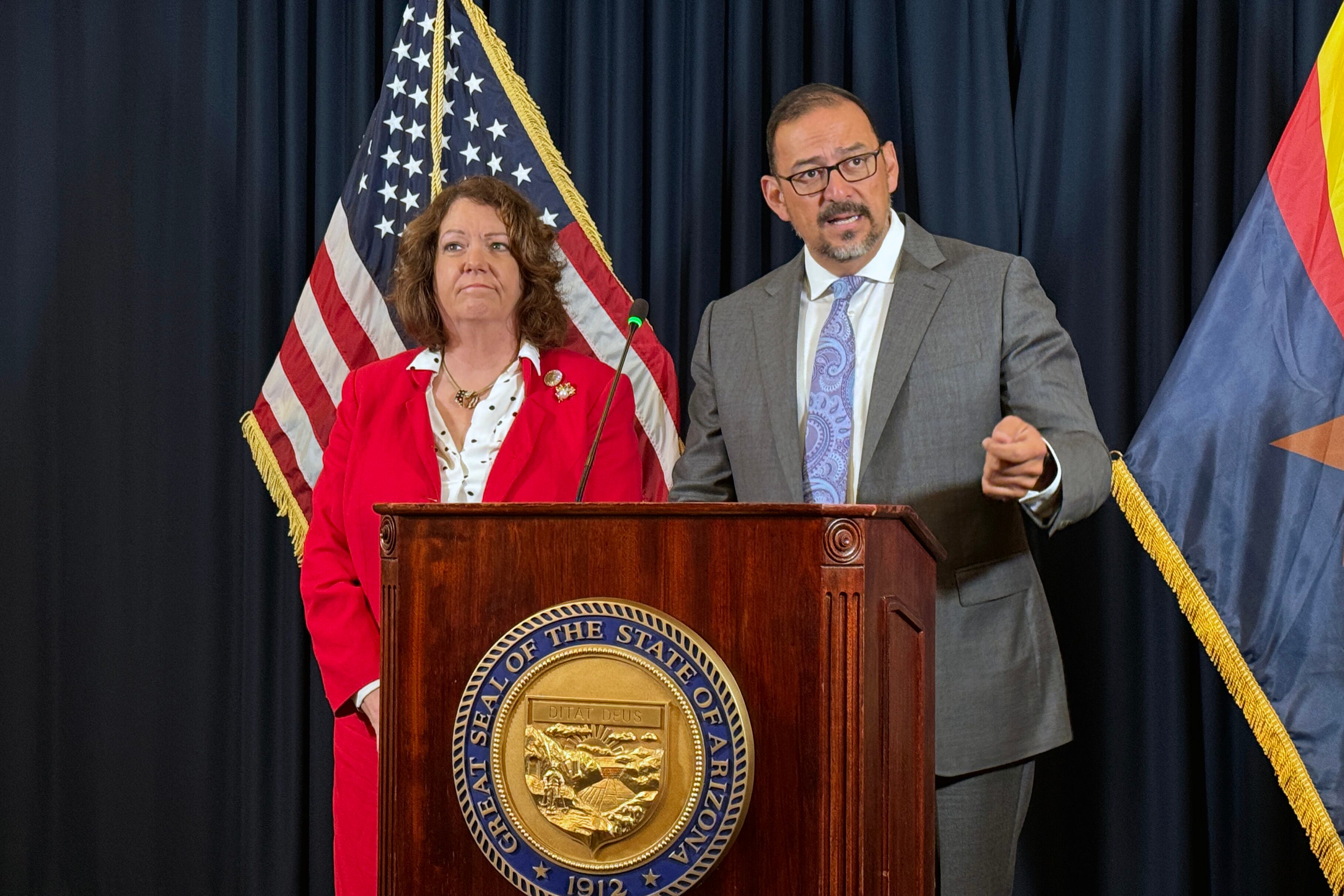 A woman and a man in business suits stand behind a wooden podium and in front of an American and Arizona flag.