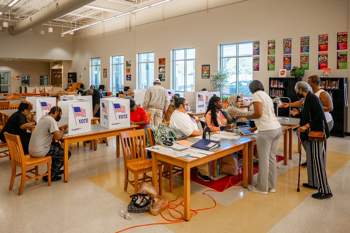 A room lined with windows is full of people sitting at tables voting.