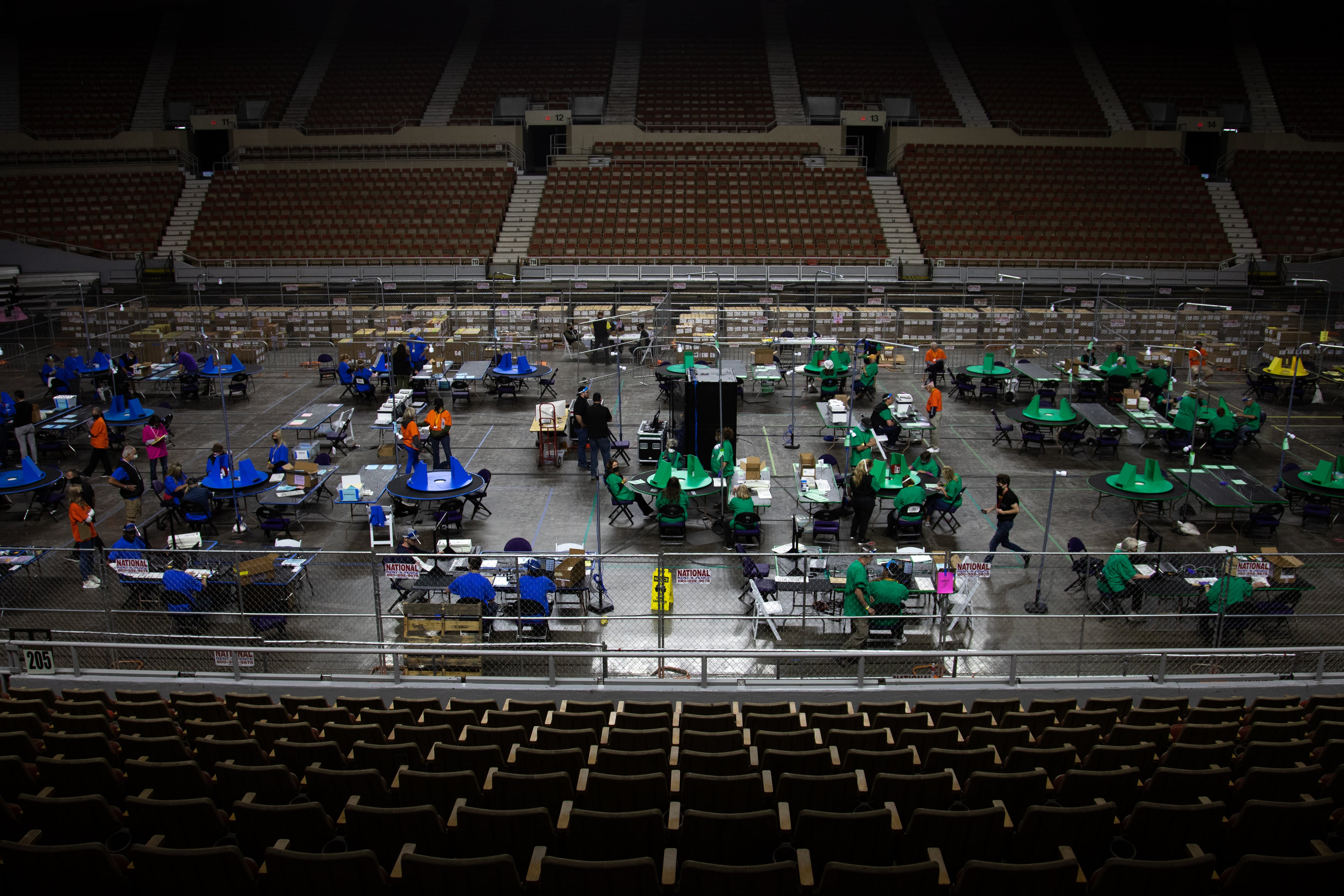 People in colored T-shirts work at several tables on the main floor of a sports arena