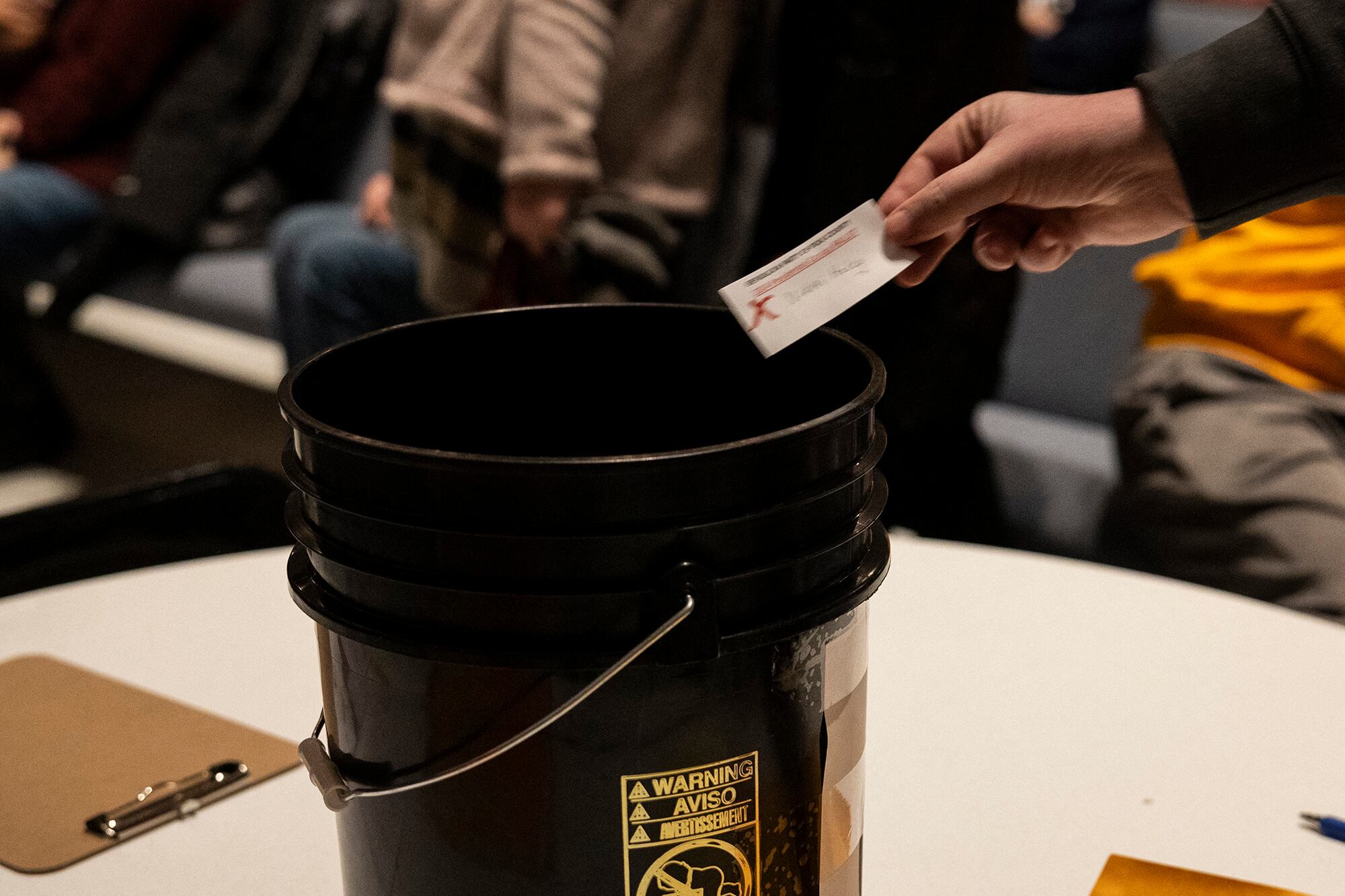A close up of a hand dropping a piece of paper into a black bucket that is on top of a table.