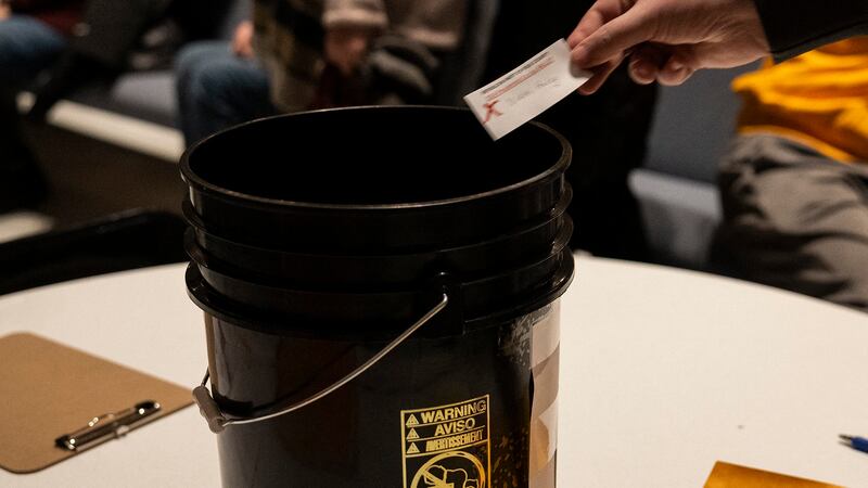 A close up of a hand dropping a piece of paper into a black bucket that is on top of a table.