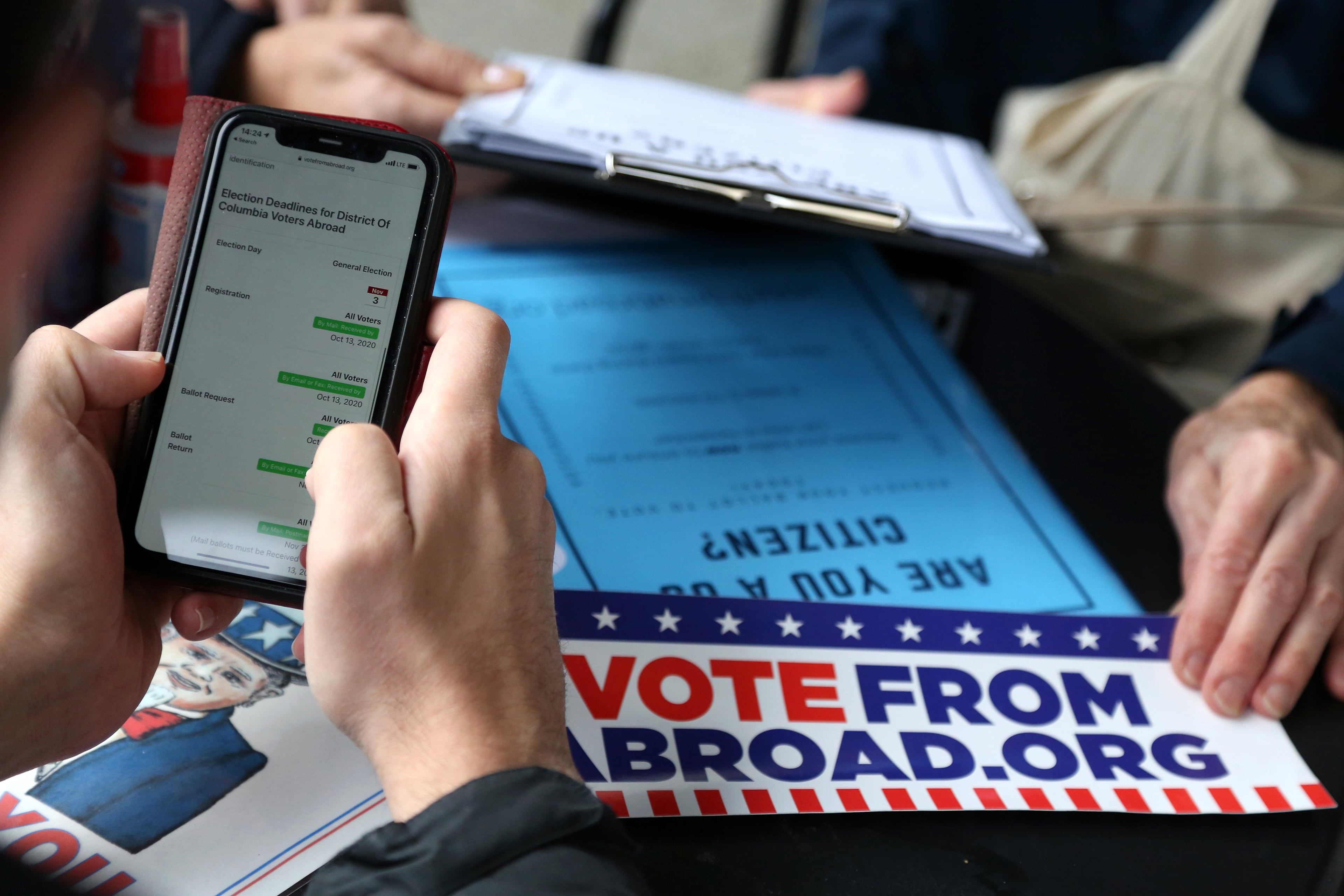 Two pairs of hands, one holding a cellphone and one filling out a piece of paper with a large sign that reads "Vote from Abroad.org."