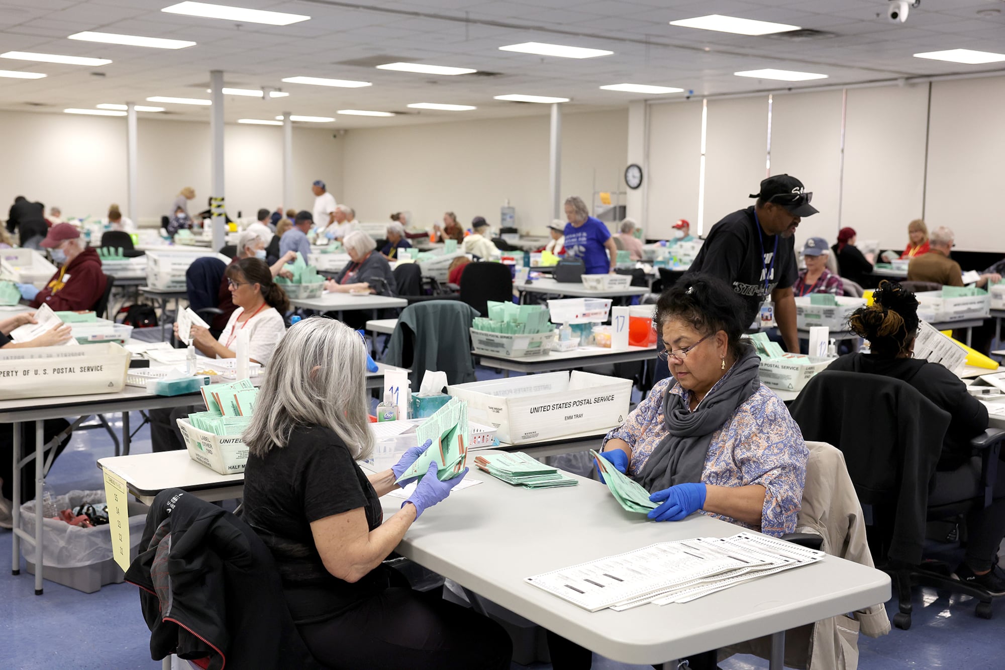 Two women sit across from one another at a table wearing blue gloves and opening ballots with other workers in the background