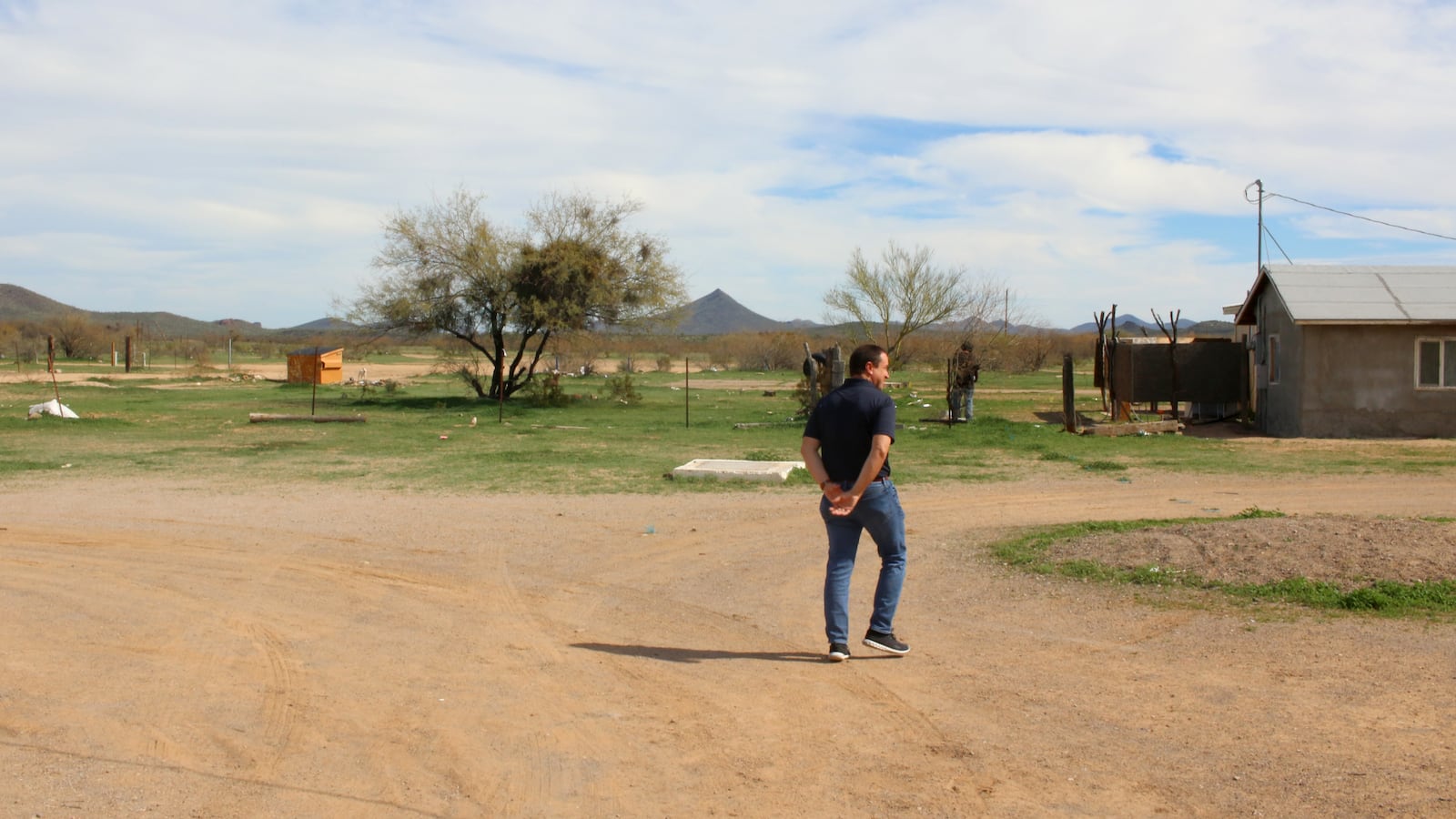 A person walks down a dirt road with a building to the right and a mountain and cloudy sky in the background.