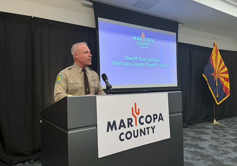 A man speaks behind a podium and in front of a projector screen and dark cloth background.