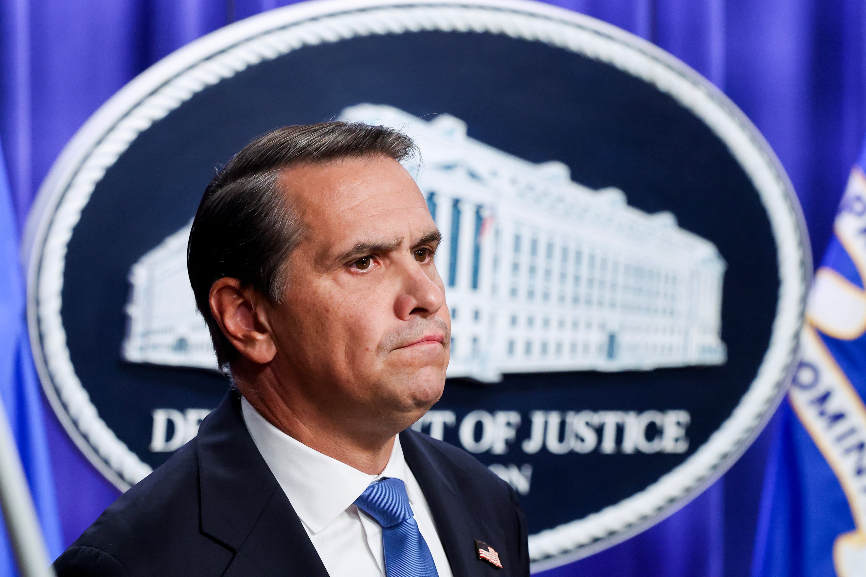 A white man in a suit stands in front of the Department of Justice logo during a press conference.