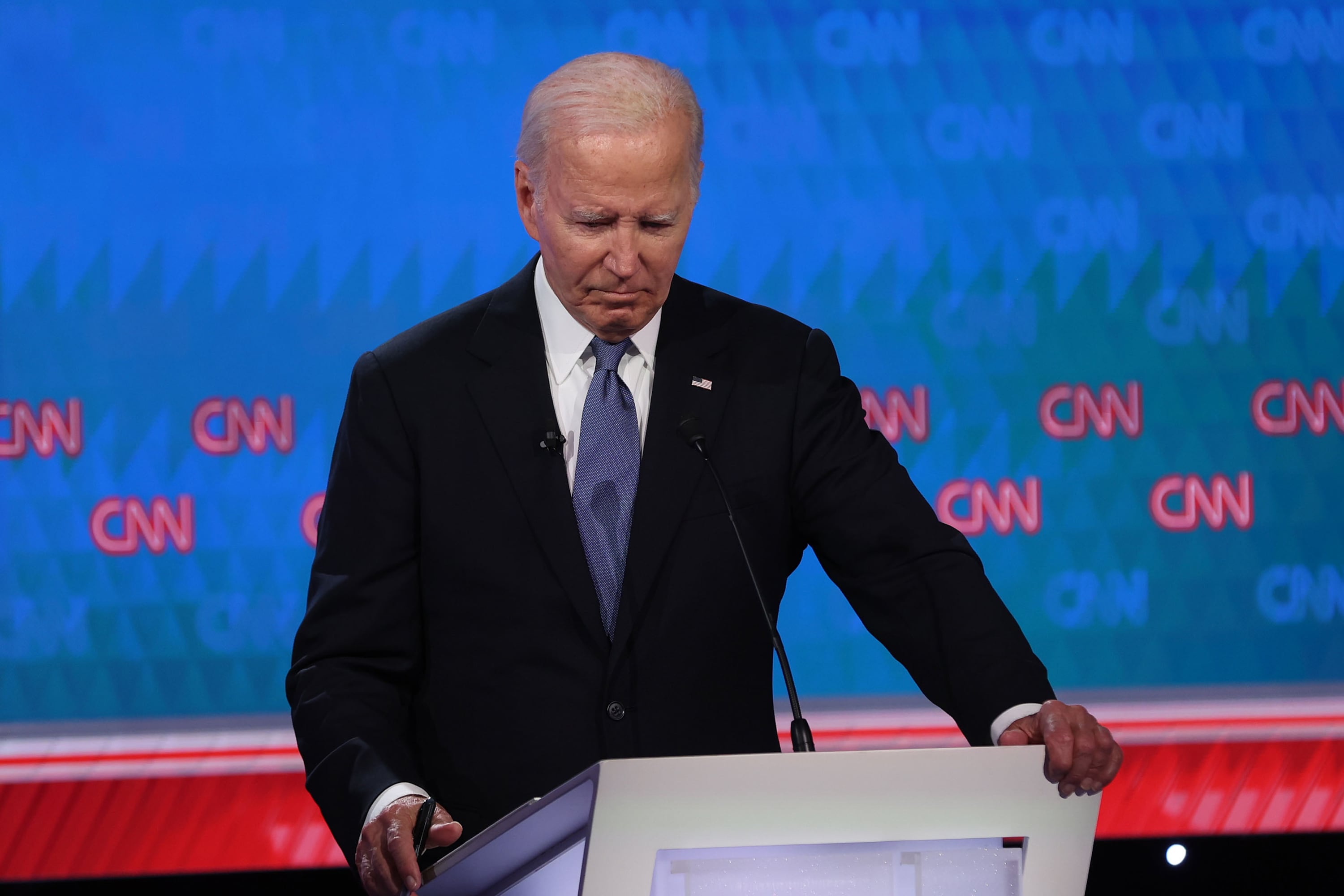 President Joe Biden wears a dark suit and blue tie and stands behind a podium with a blue and red screen that says CNN in the background.