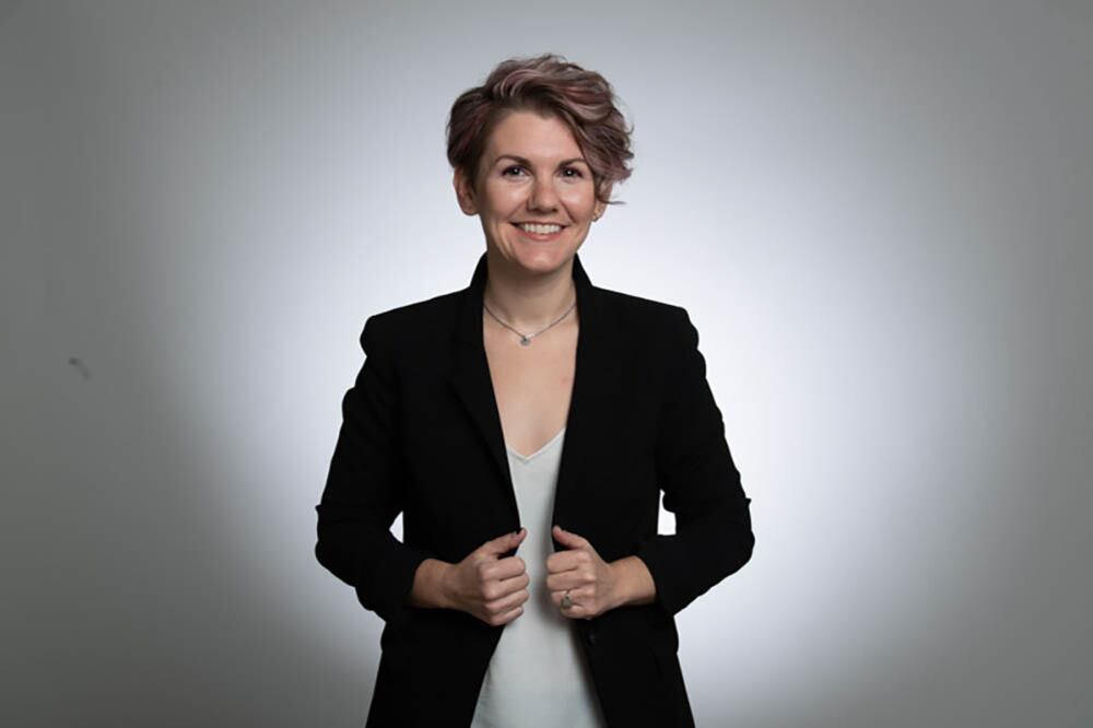 A woman with short brown hair and wearing a dark suit jacket smiles for a portrait in front of a light background.