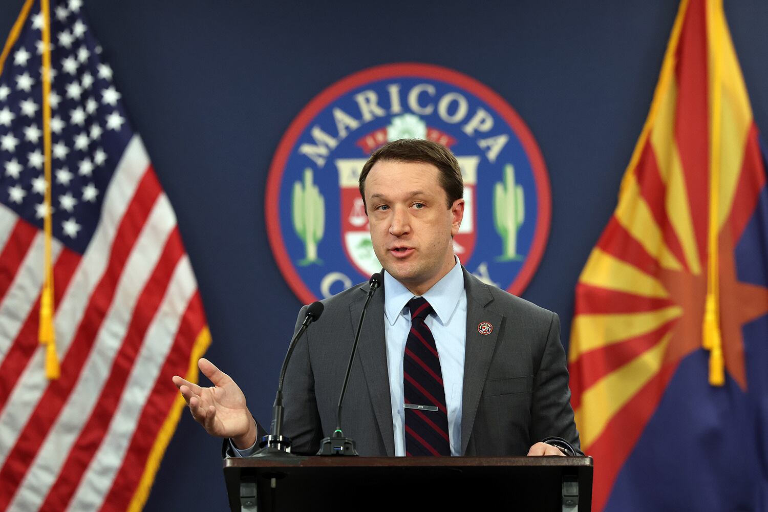 A man with short dark hair and wearing a suit stands at a podium with the Maricopa seal and American and Arizona flags in the background.