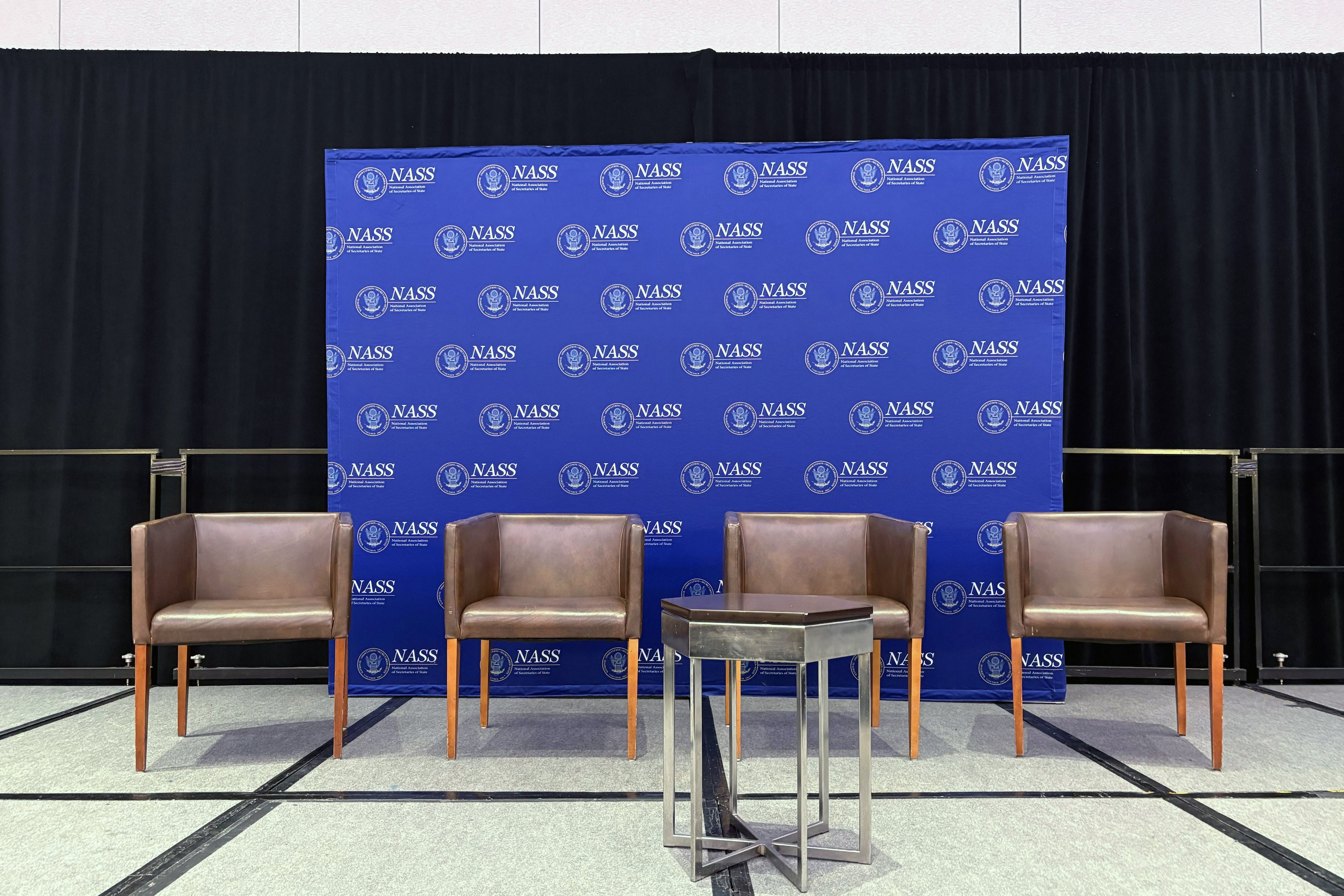 A photograph of four empty chairs on stage with a blue and white logo banner in the background.