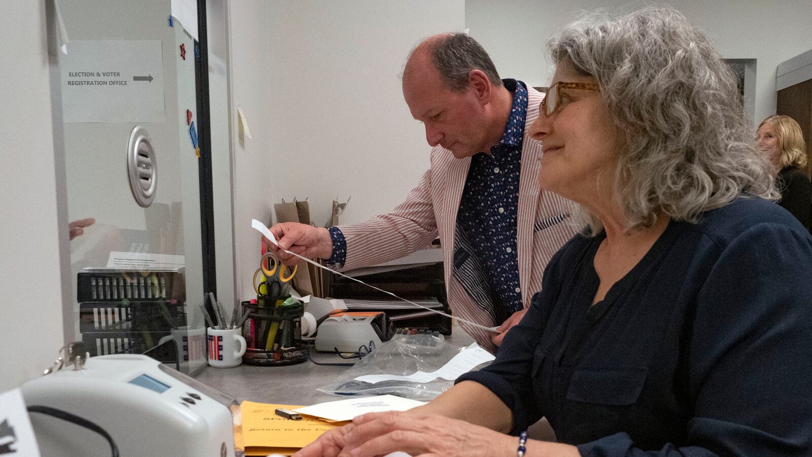 Two people stand and work from a desk with papers and various things.