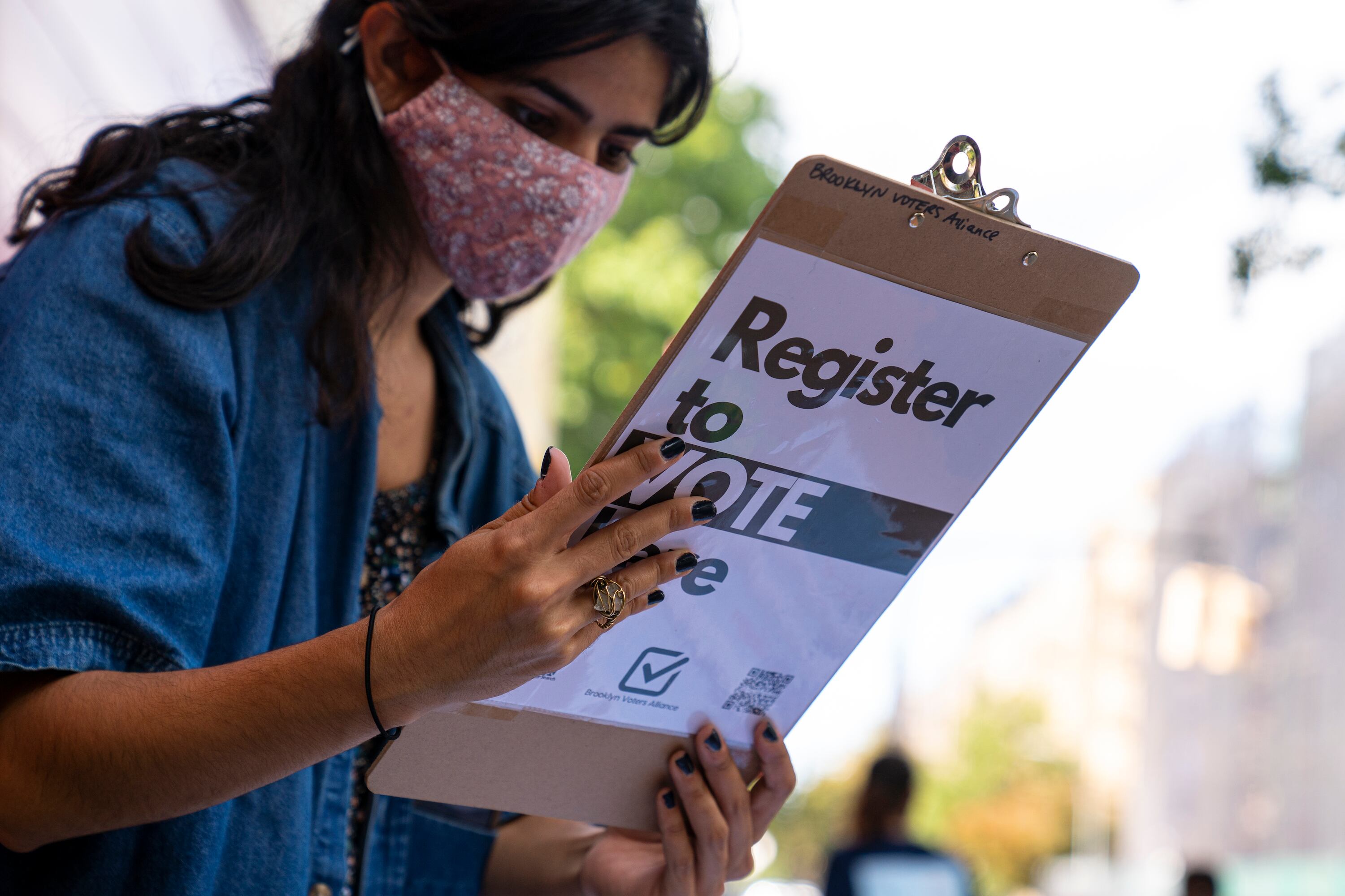 A woman with long brown hair wearing a face mask checks something on a clipboard that says “Register to Vote” on the back of it.