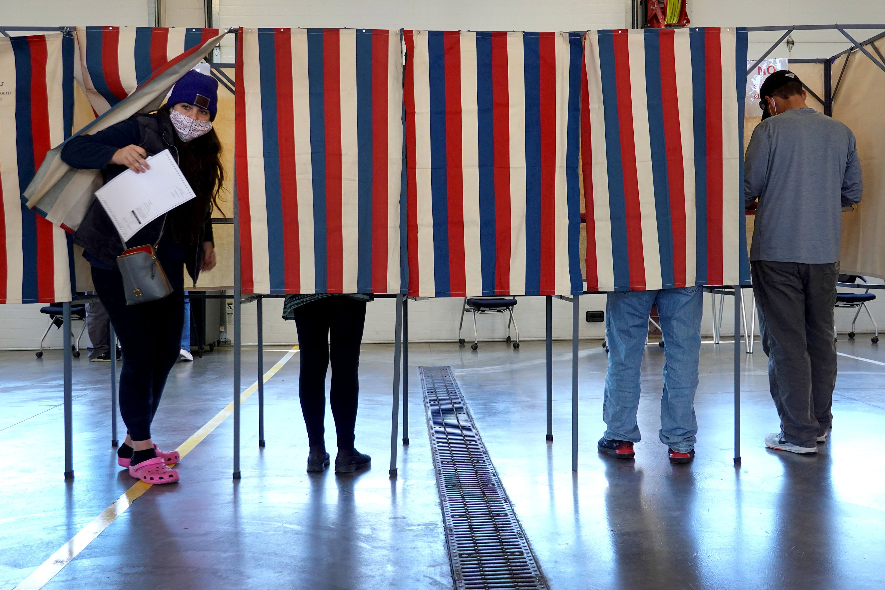 A person holding a ballot walks out from behind a voting booth curtain while three other people stand in the booth voting.