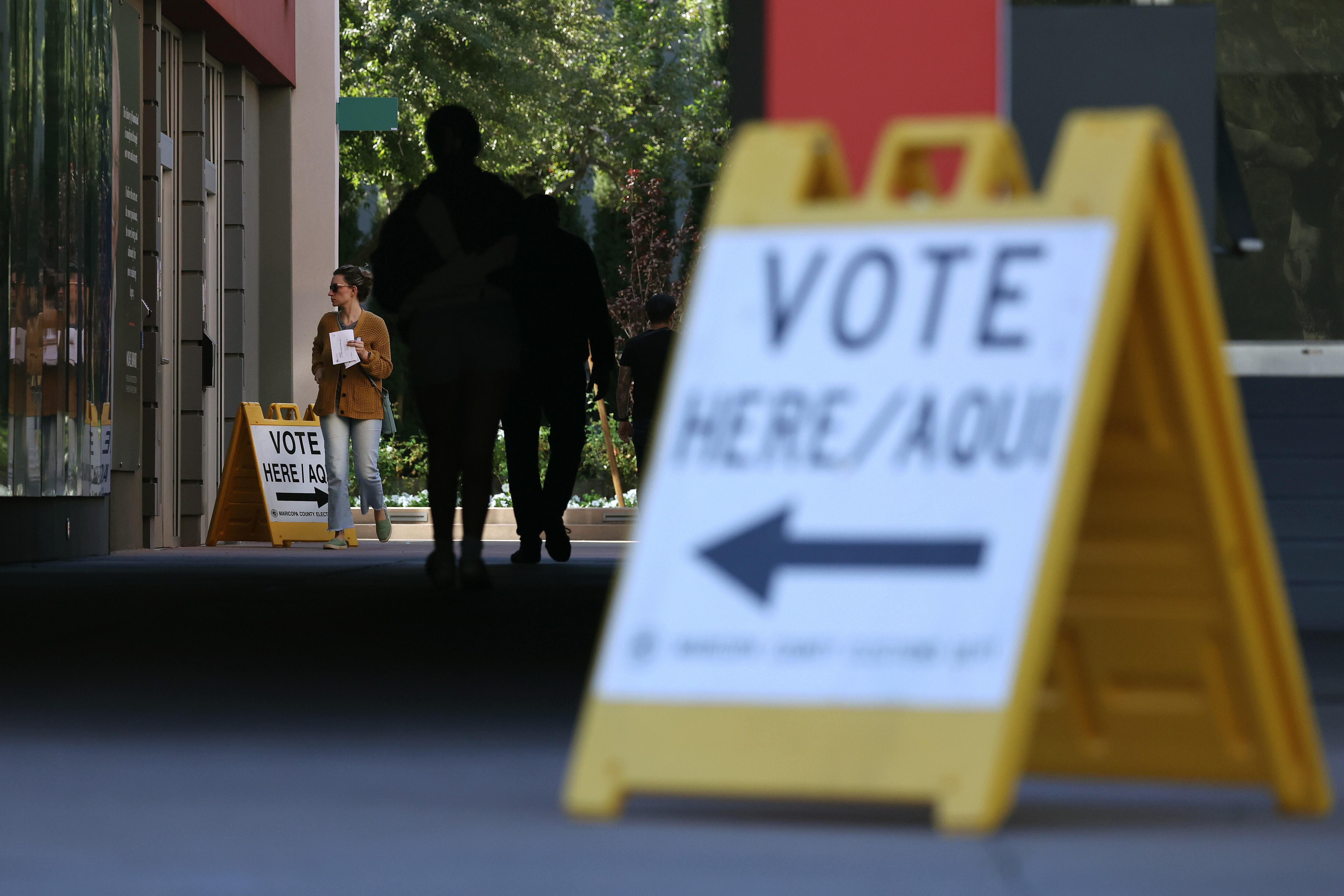 A large black and white sign that reads "VOTE HERE/AQUI" with people walking in the background.