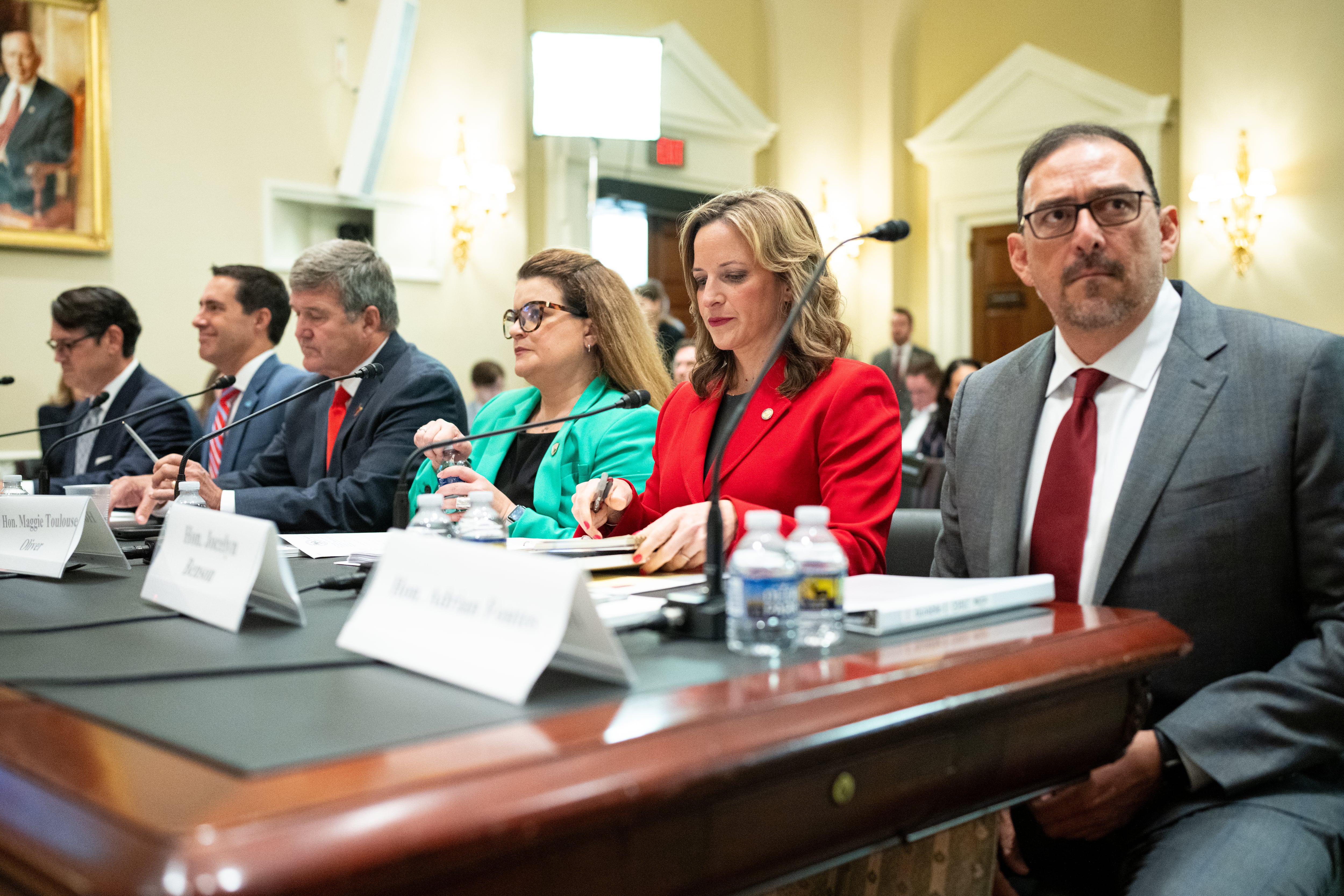 A photograph of six people in business clothes sitting side by side at a long wooden table with microphones, name tags and notebooks in front of them all in a large room.