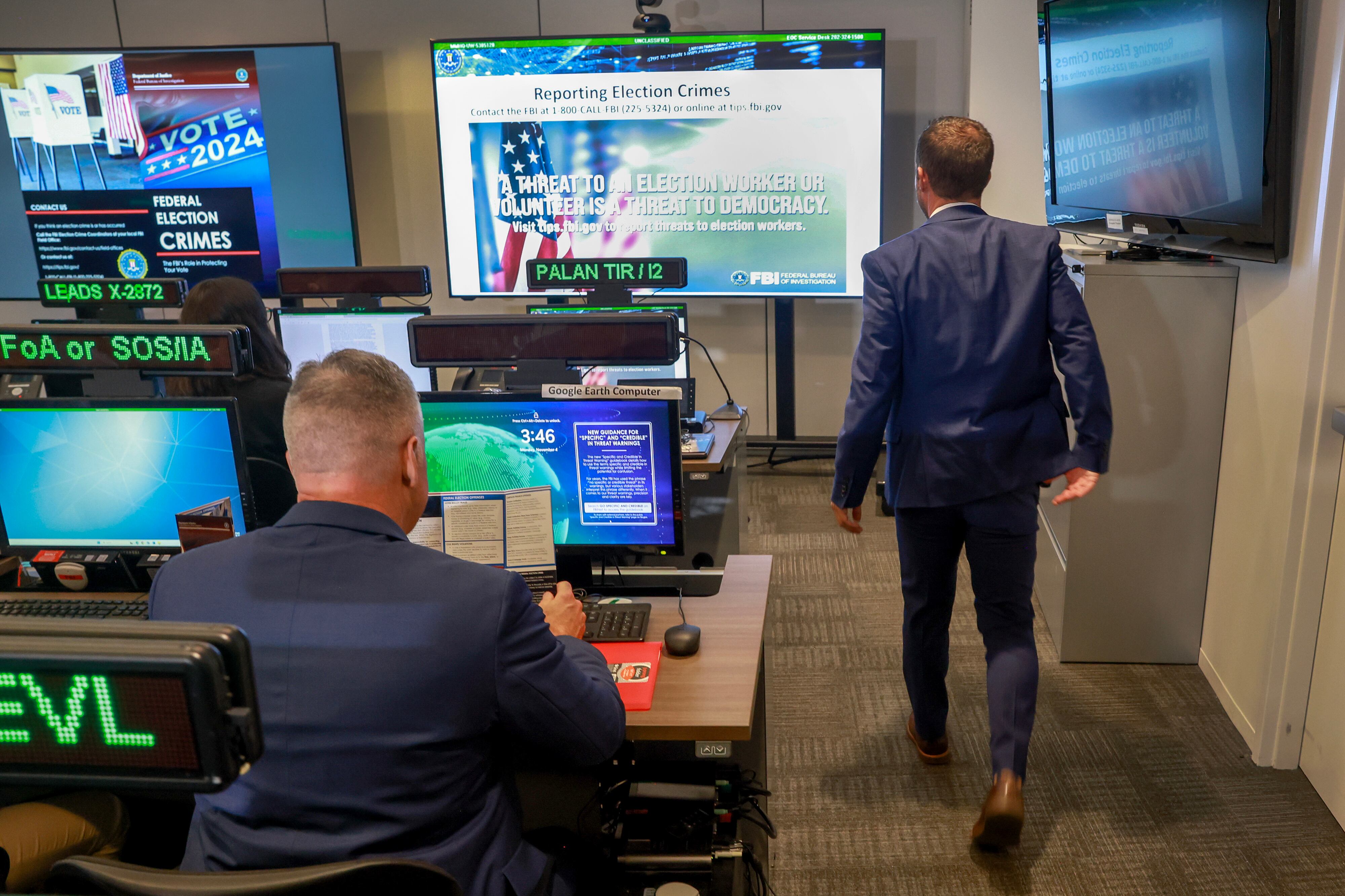 A man walks by two rows of computers with one person sitting in the foreground while three large tvs line the wall on the right and the back wall.