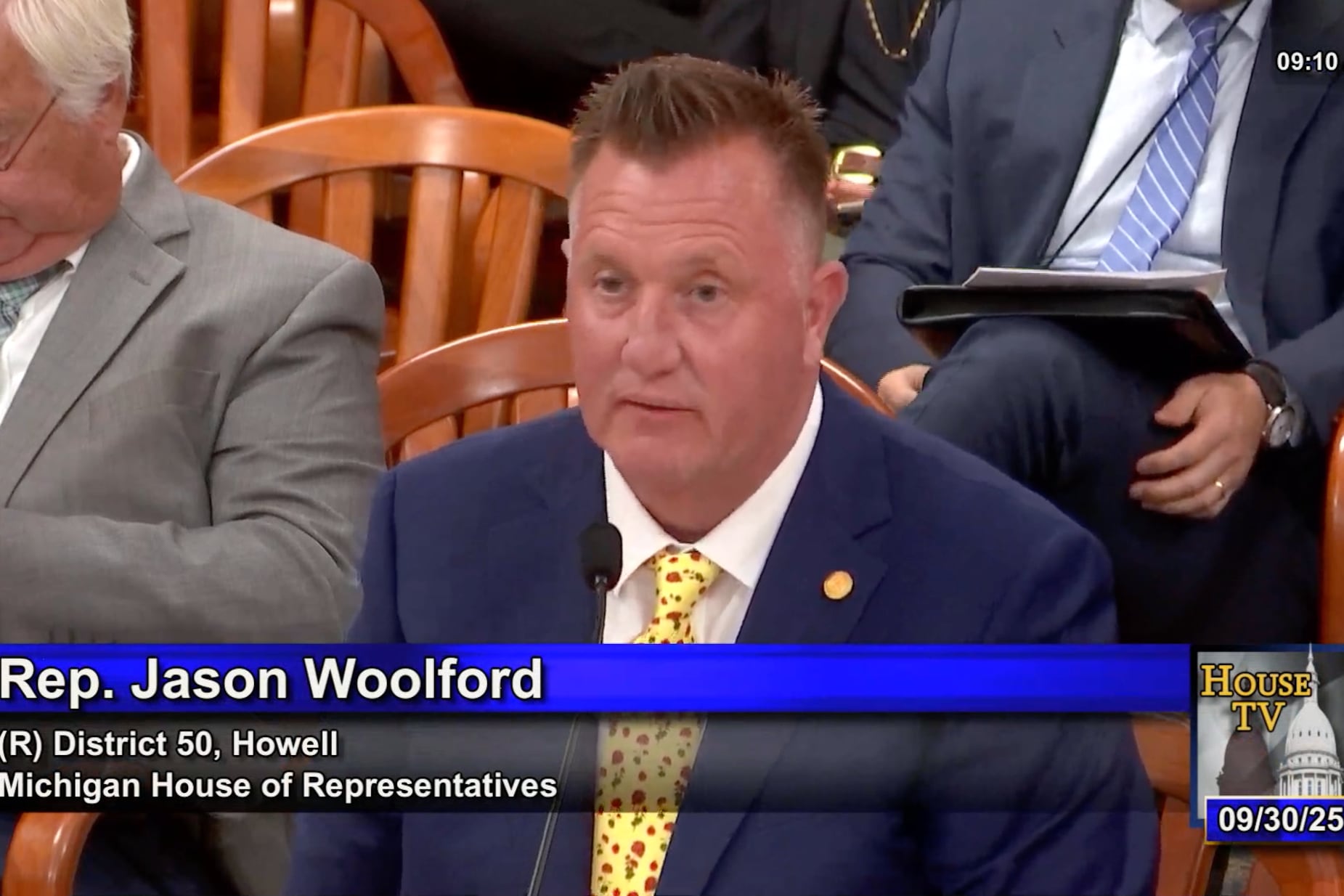 A screen grab of a white man in a suit sitting in front of two other men in suits.
