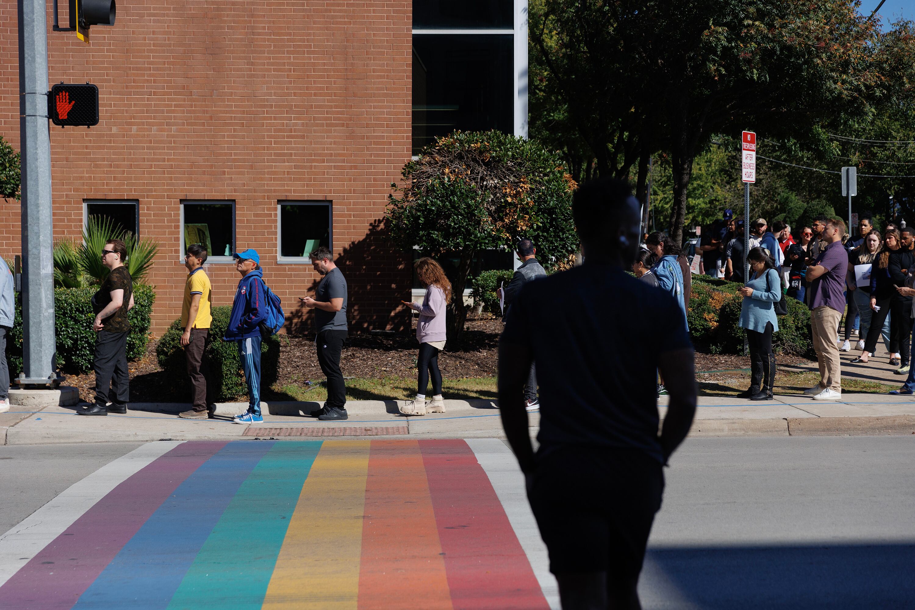 A long line of people stand outside with one person walking toward the line of people in the foreground.