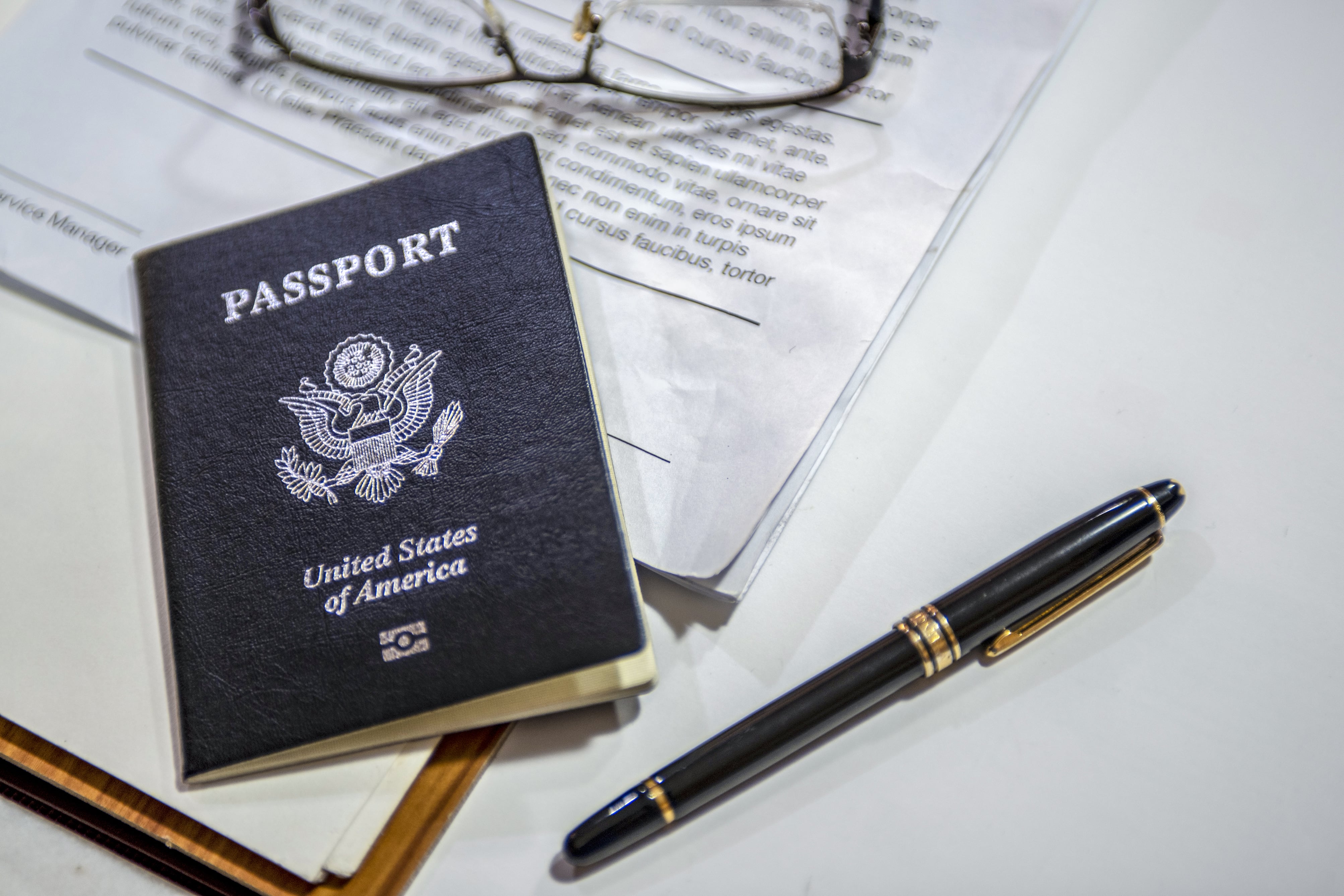A blue and white US passport on a desk on top of papers next to a pair of eye glasses and a pen.